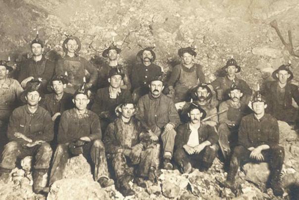 historic sepia photo of a group of men in mining gear, they are in the mine siting on a pile of rock.