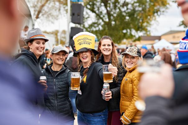 a group of woman at an outside festival, pose for a photo. They are holding mugs of beer