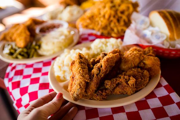 A woman's hand grabs a plate of fried chicken and sides off a table with a red and white checkered table cloth. In the background, there are additional plates of fried chicken.