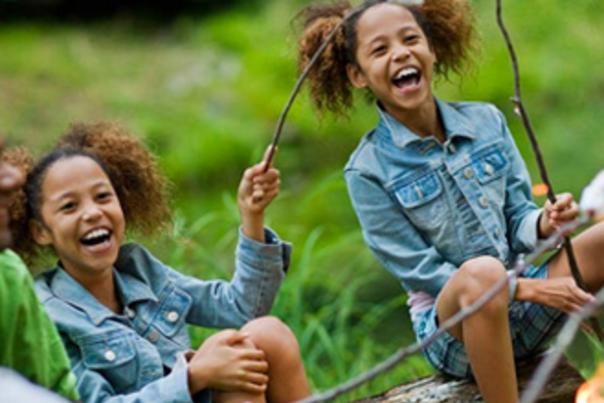 Three young people sitting round a campfire toasting marshmallows on sticks, two are dressed in similar clothes with similar hairstyles, perhaps twins?