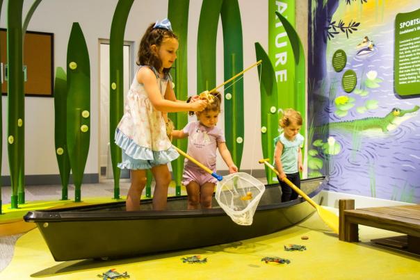 three children play in a boat and pretend to fish at the children's museum