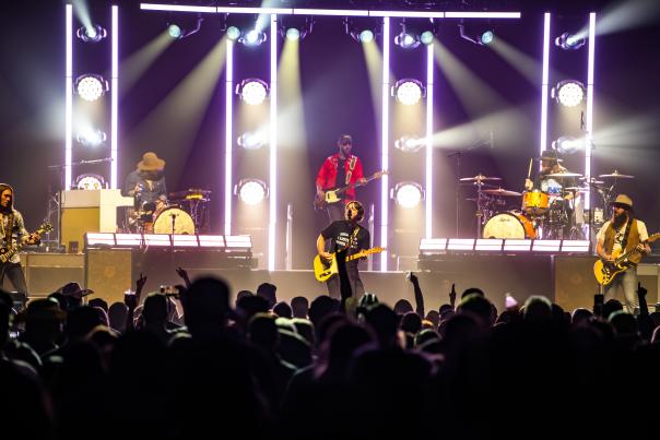 A live music concert featuring a full band performing on stage, with dramatic vertical light bars and colorful spotlights illuminating the scene. The lead singer stands center stage with a yellow electric guitar, flanked by guitarists, a bassist in a red shirt, and two drummers. The crowd in the foreground is silhouetted, many raising hands and taking photos, contributing to the vibrant, high-energy atmosphere of the show.