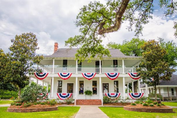 two story white historic home and museum with red, white, and, blue flags draped on the balcony, and trees