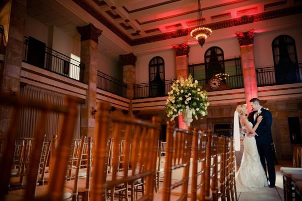 couple poses in wedding gown and tux beneath a chandelier in a multi-story building