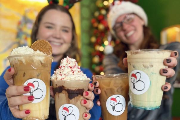 two women in a santa hat and reindeer ears hold four frozen and iced coffees. One is topped with whipped cream and a cookie, the next with whipped cream and red sprinkles, the next is a darker brown, the last has a blue drizzle at the bottom.