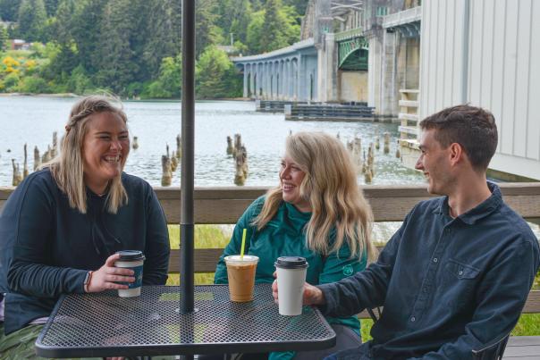 Three people with coffee drinks sit at a bistro table with the river and bridge behind them.