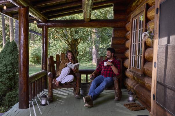Two people sit on the porch of the log lodge. One reads a book, the other sips a warm beverage.