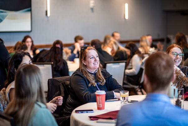 A room full of people sitting at round tables during a meeting. The focal point is one woman smiling across the table, with nearby people smiling and talking.