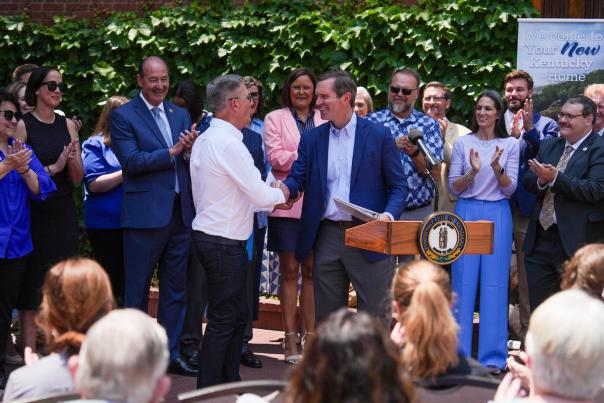 Press conference with two men shaking hands at podium in front of a crowd of people