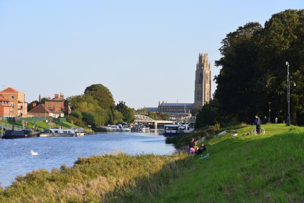 A cycle path srurronded by grass and trees next to a river. Boston Stump and town can be seen in the distance.