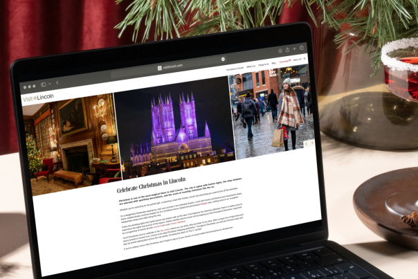 An open laptop showcasing a Christmas page, with a red cocktail nearby and small Christmas tree on the table behind the laptop.