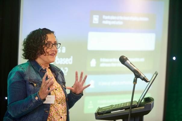 A woman presenting in front of a microphone and laptop, with a large screen behind her.