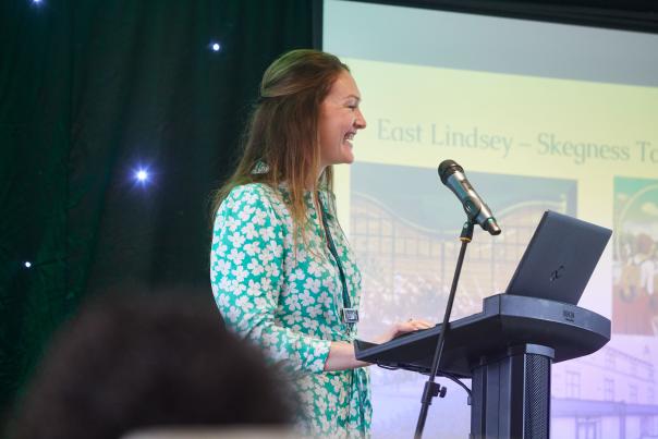 A woman stood at a speaker podium with a microphone smiling with presentation slides in the background.