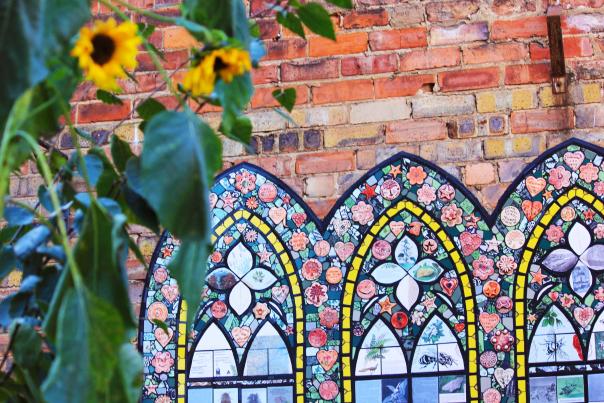 An art installation in the style of a stone glass window on a brick wall behind some sunflowers.