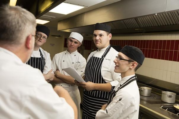 A group of culinary students listening to instructions in a restaurant kitchen.