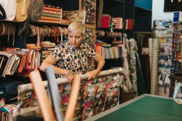 A woman rolling a long piece of fabric in a shop, with a wall full of fabrics behind her.