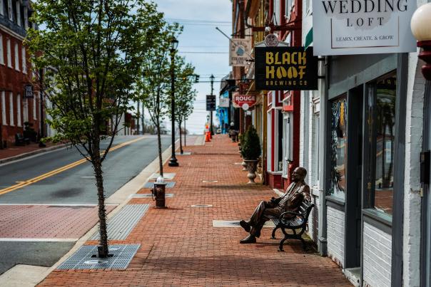 Bronze statue seated on a bench along the brick sidewalk of downtown Leesburg, with Black Hoof Brewing Co. and historic storefronts visible along King Street