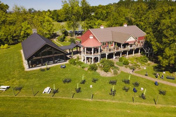 Aerial view of Cana Vineyards. A large, rustic building with a red roof and a modern black structure nearby, surrounded by green lawns and trees.