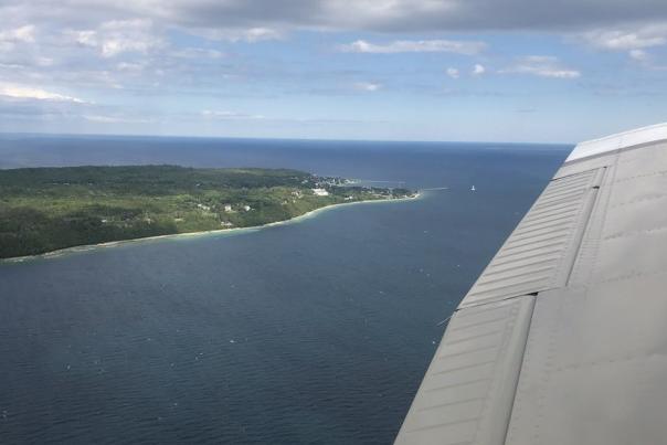 Green trees of Mackinac Island and surrounding blue water seen from above with the wing of an airplane in the foreground