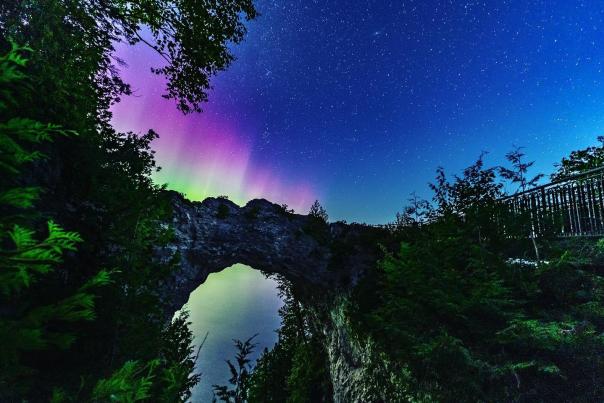 Red and green colors of the Northern Lights emerge in the night sky behind Arch Rock on Mackinac Island