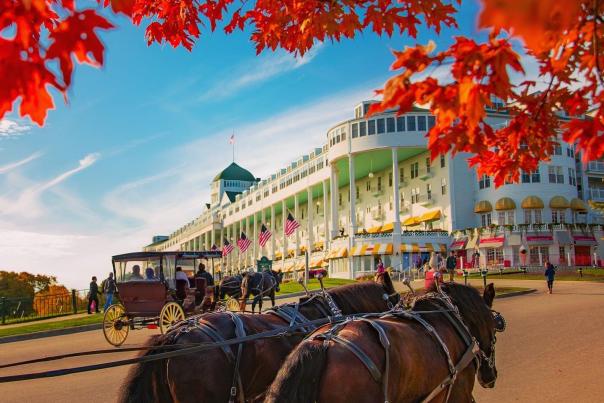 A team of horses pulls up toward Mackinac Island’s Grand Hotel in fall with orange leaves on the trees.