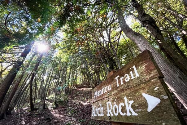 The sun peeks through the trees in Mackinac Island State Park above a sign pointing to Arch Rock and Manitou Trail