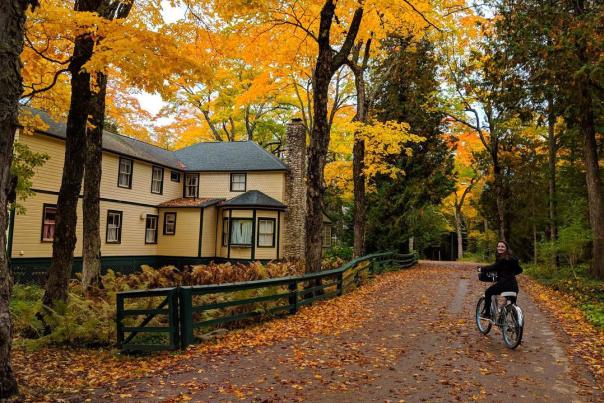 A woman pedals a bicycle on a Mackinac Island road filled with leaves past a yellow cottage and trees full of fall color