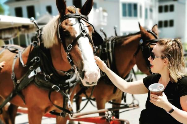 A Mackinac Island visitor holding a beverage in one hand pets a team of horses with the other hand on a sunny day