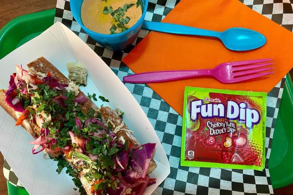 A view from above of an open-face sandwich, a side of yellow soup, Fun Dip and brightly colored cutlery