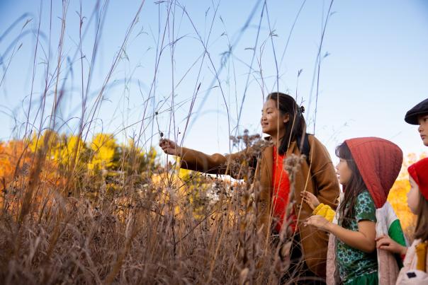 A woman touches a piece of fall foliage at a park with three small children nearby