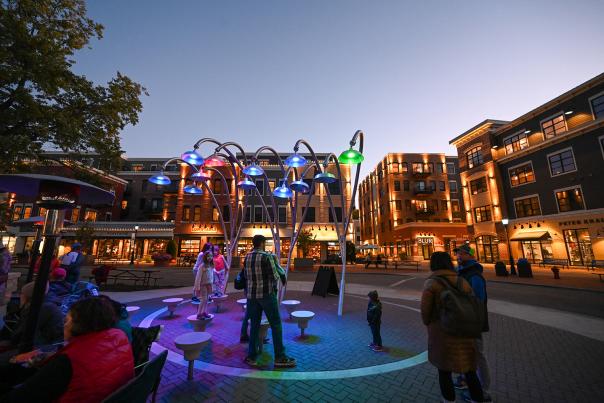 Colorful light fixtures illuminate a lively plaza at dusk, with people interacting and enjoying the vibrant atmosphere near shops and restaurants.