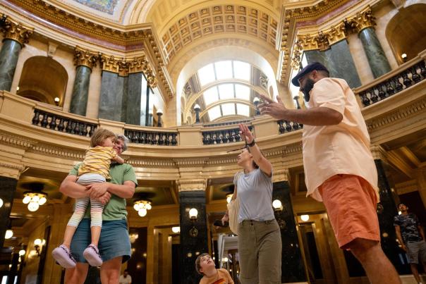 A family explores a grand, historic building with ornate architecture. They engage in conversation, surrounded by marble columns and large windows.
