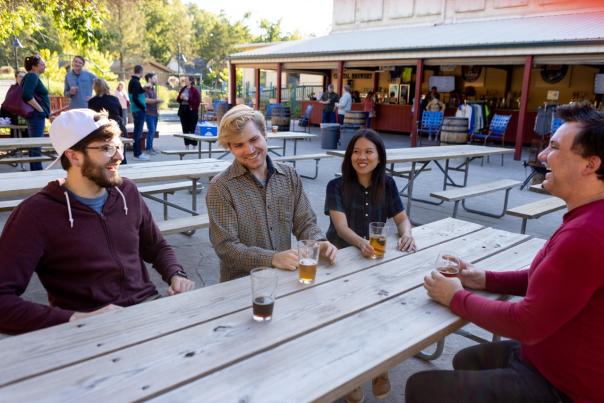 Four friends sit at a wooden picnic table outdoors, enjoying drinks in a lively setting with people and a bar in the background.