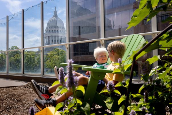 Two young white children sit on a char surrounded by plants on the Madison CHildren's Museum rooftop with the Capitol building in the background.
