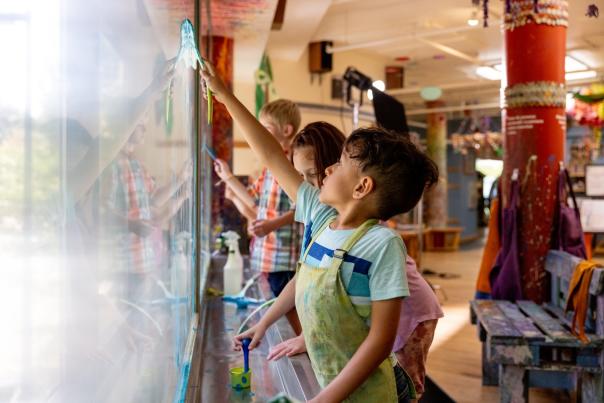 A boy reaches up to paint on a window in the art studio at the Madison Children's Museum. Two other children are next to him also painting.