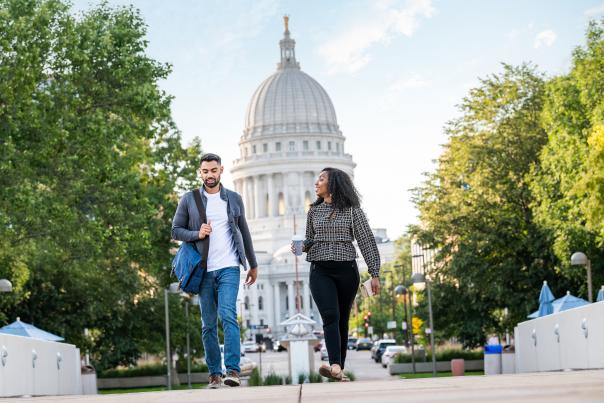 A man and woman walk away from the Wisconsin State Capitol toward the Monona Terrace. The main has a laptop bag over his shoulder and the woman is carrying a cup of coffee.