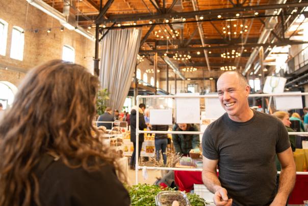 A white man smiling while working a booth at an indoor farmers' market.