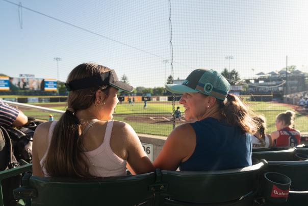 Two people sit in stadium seats, talking while watching a softball game on a sunlit field behind a protective net.