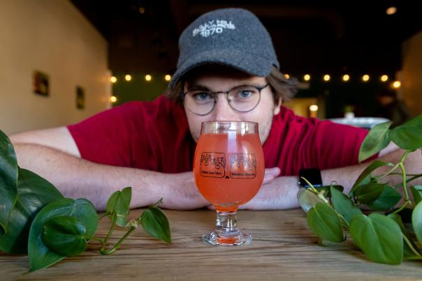 A white man peeks from behind a glass of cider on a table surrounded by plants at Hidden Cave Cidery