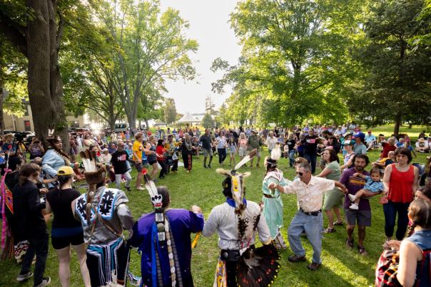 A group of dozens of people, some in Ho-Chunk attire and some not, gather in a circle and dance and sing at San Damiano Park