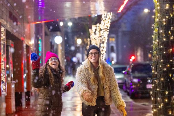 Two women walking and throwing snowballs in downtown Madison as the trees are lit up with holiday lights.