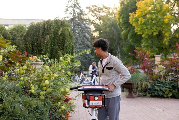 A young Black man wheels a bicycle through lush greenery at Allen Centennial Gardens