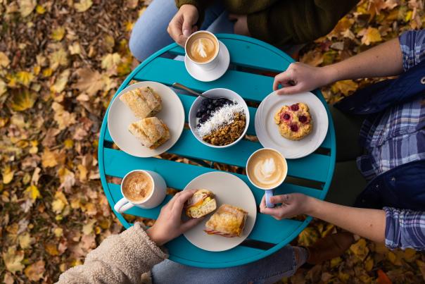 An overhead shot of food and coffee at a table outdoors with leaves on the ground underneath.