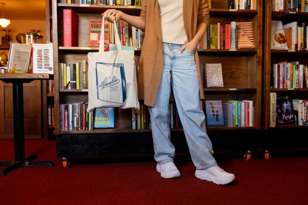 A photo from the chest down of a person holding a Leopolds Bookstore tote bag while standing in front of a bookshelf.