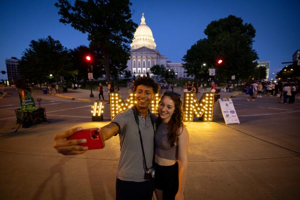 Two people take a selfie in front of the light up MNM sign in front of the Capitol building