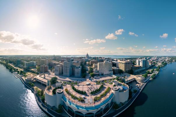 Aerial view of a vibrant city skyline along a river, featuring modern buildings, green spaces, and a clear blue sky. Sunlight reflects on the water.