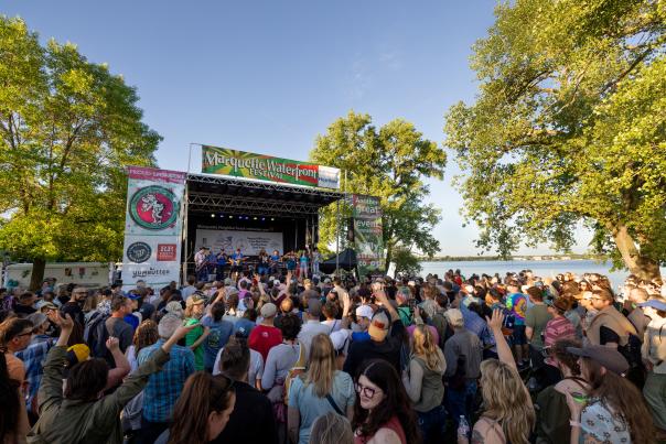 A lively outdoor music festival scene with a large crowd in front of a stage, surrounded by trees and a waterfront view. Bright banners promote the Marquette Waterfront Festival..