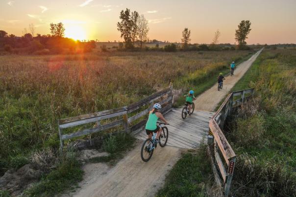 Four bikers cross a wooden bridge surrounded by green pastures. The sun is setting in the distance.