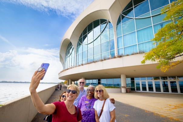 Group of five people taking a selfie in front of a modern building by a waterfront, with a clear sky and trees nearby.