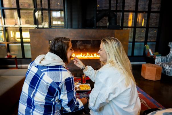 Two white women embrace in front of a fireplace at the Moxy hotel. One woman feeds cheese to the other woman.